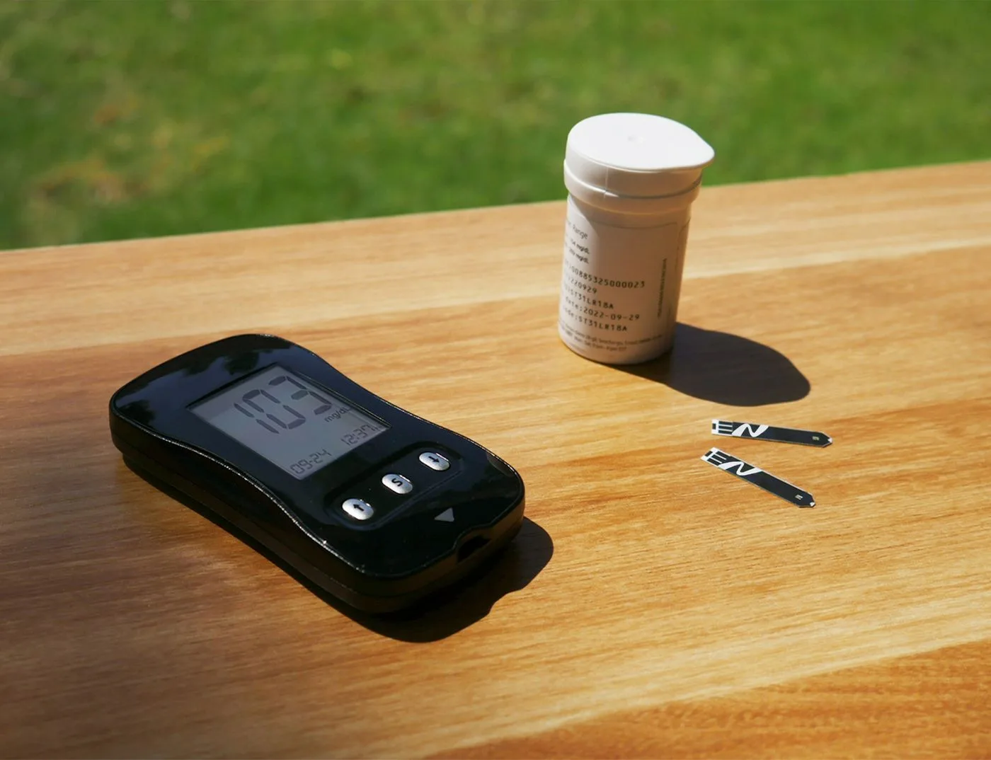 A glucose meter and unused testing strips on a sunlit wooden table.